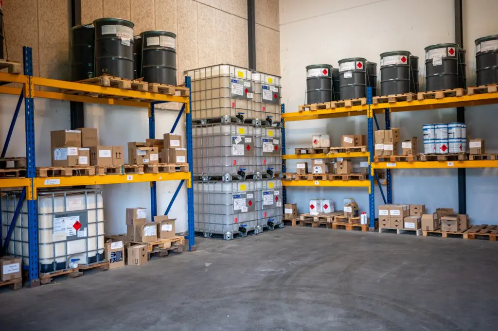 Lagerrum med hylder fyldt med tønder, IBC-beholdere og kasser med kemikalier, klar til logistik og distribution.