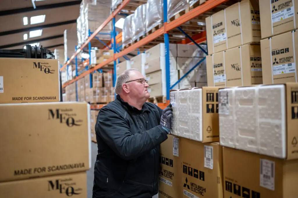Lagerarbejder i handsker tager fat i en kasse med MITSUBISHI ELECTRIC mærke, mens kasser er stablet højt i et moderne lagerfacilitet.