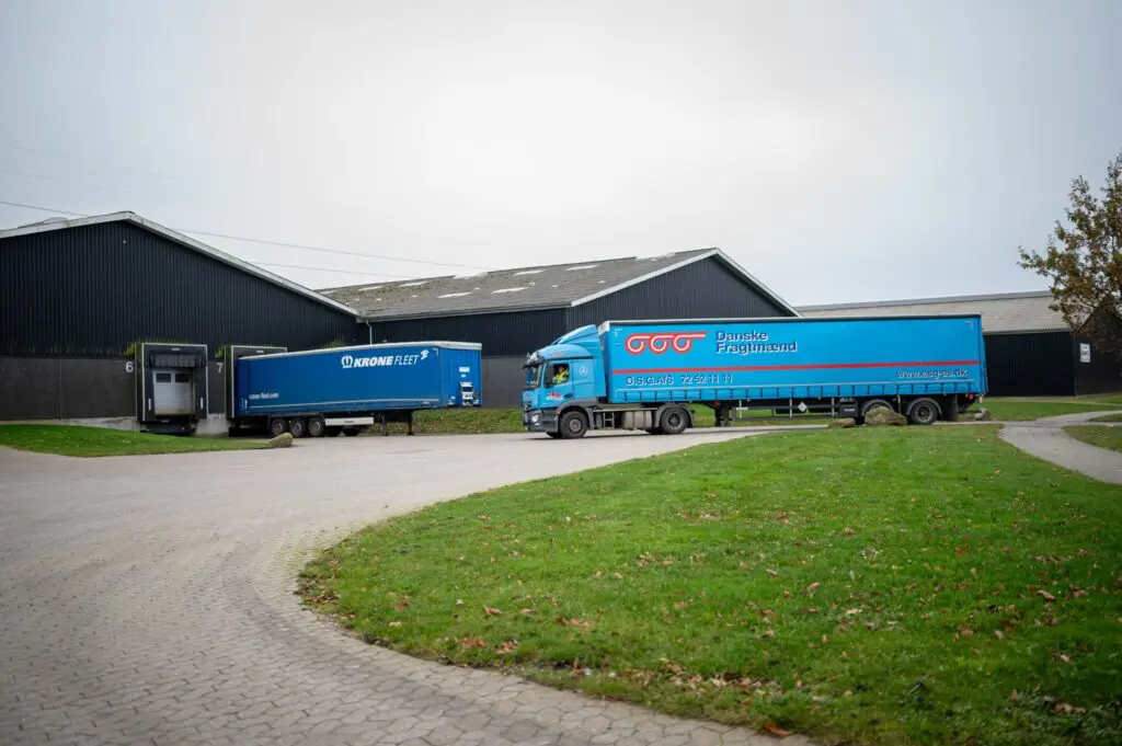 En blå lastbil med Danske Fragtmænd-trailer kører forbi en lagerbygning, hvor en Krone Fleet-trailer holder parkeret ved en læsserampe.