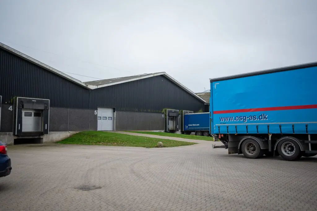 Blå lastbil med 'www.osg-as.dk' på siden ved et stort lager til logistik og distribution.
