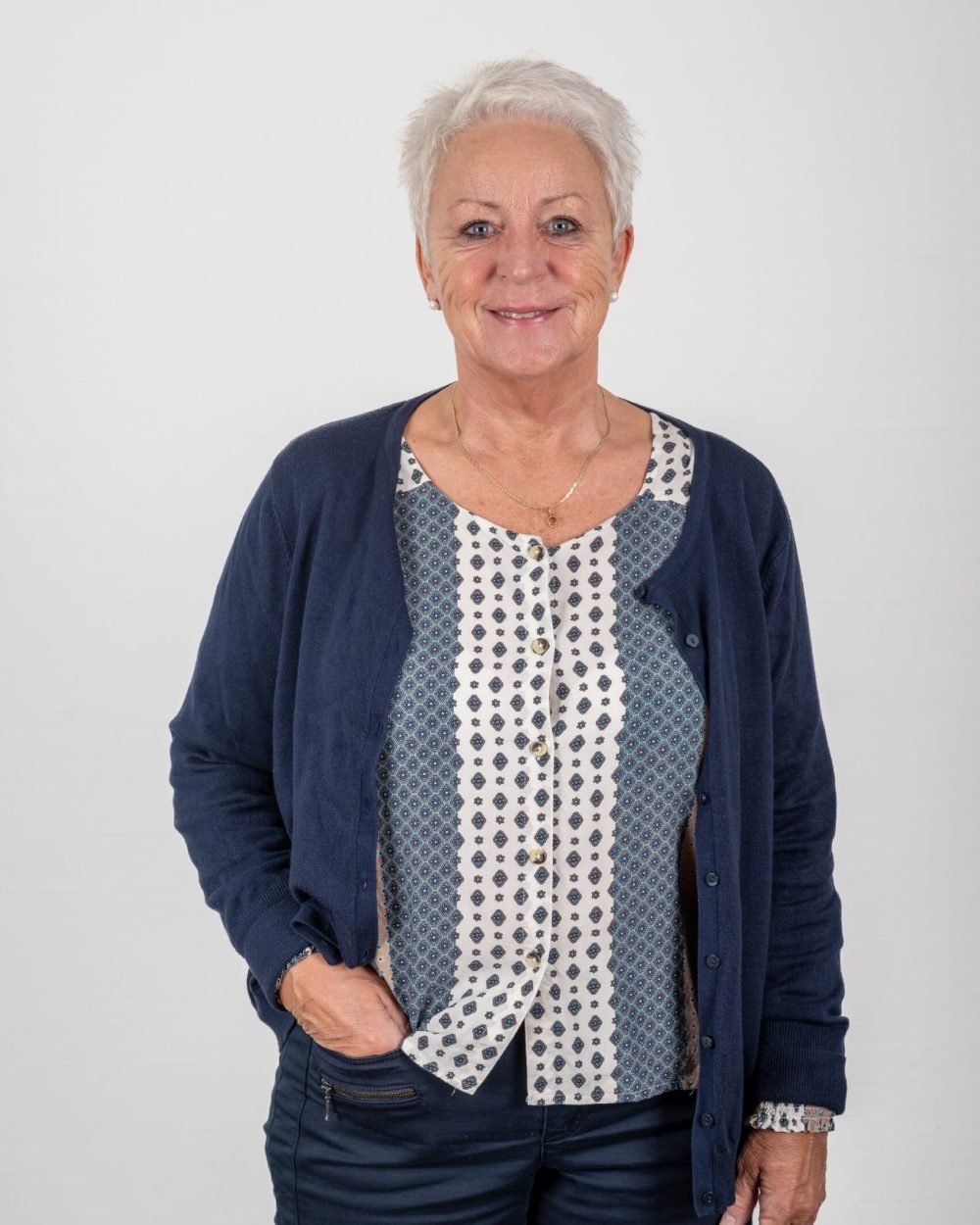 Glad kvinde med gråt hår og blå øjne, iført mørkeblå cardigan og mønstret bluse.