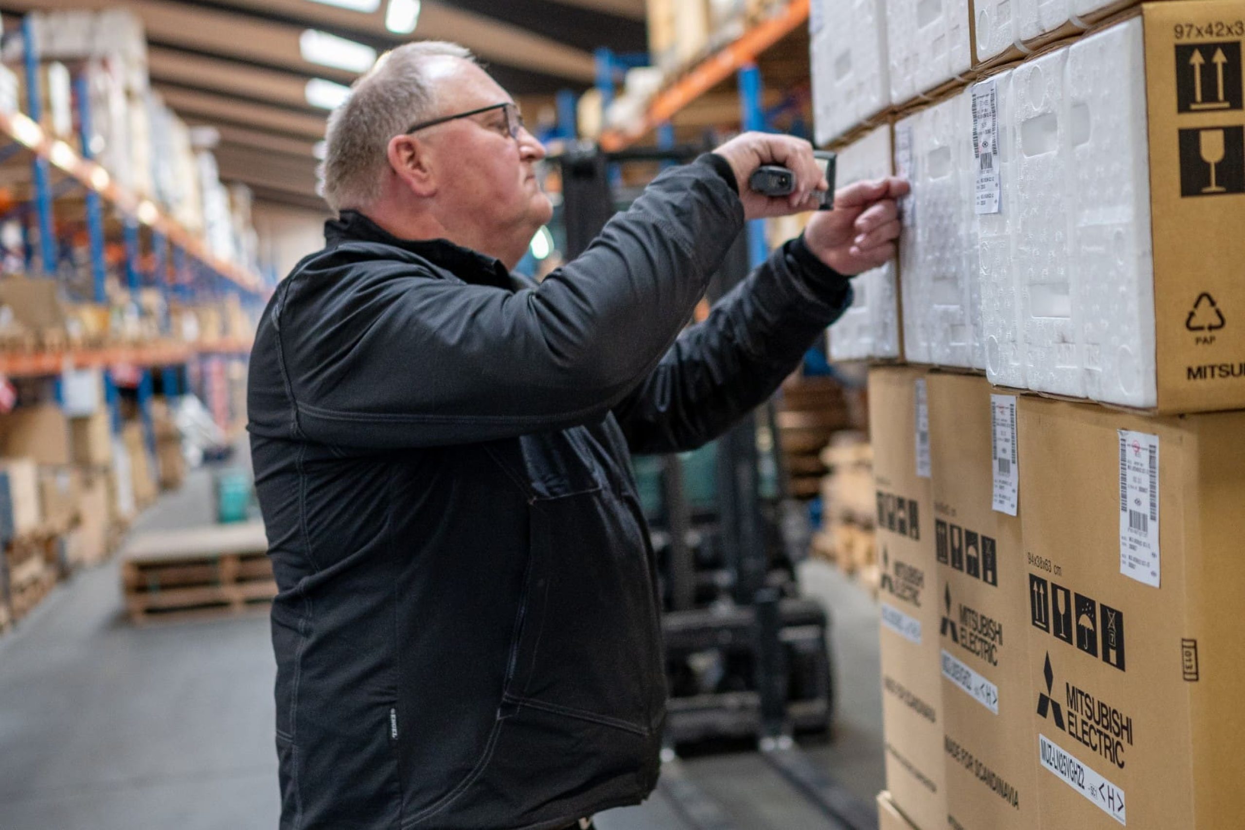 Lagerarbejder scanner stregkode på en papkasse med Mitsubishi Electric produkter i et moderne lagerfacilitet hos Behrens Logistik.